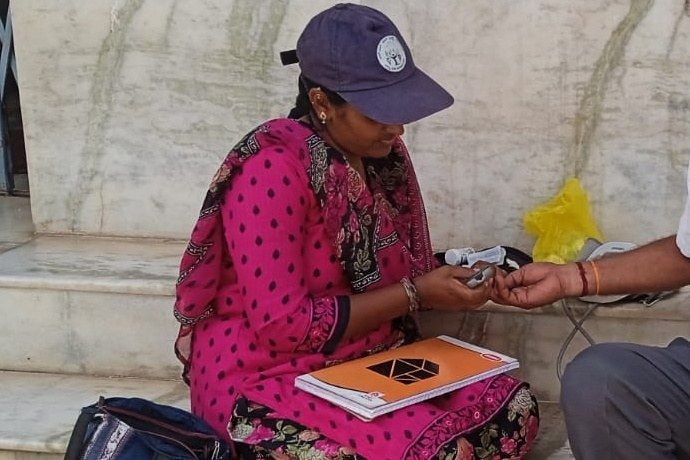 A health worker in a cap checks a resident's blood sugar with a handheld glucometer during a Parivarthana doorstep screening.
