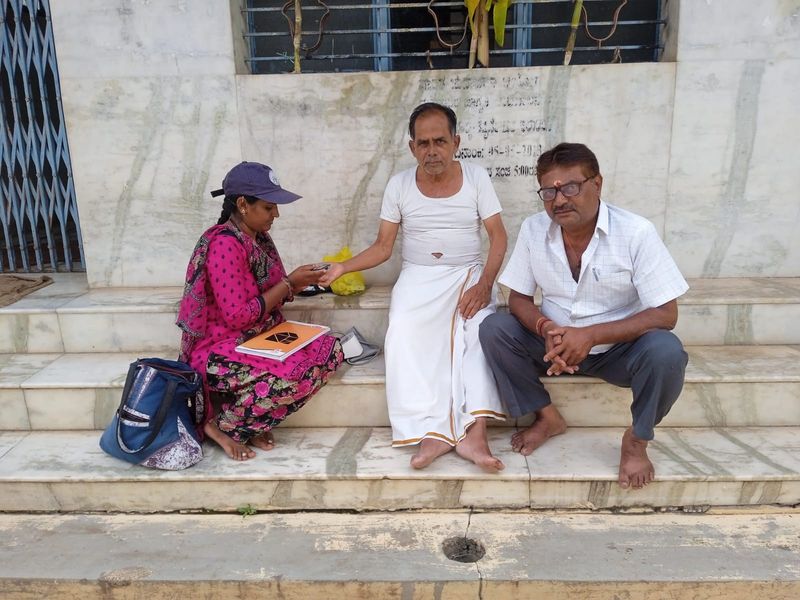 A health worker screens two elderly men seated on the steps of a village building.