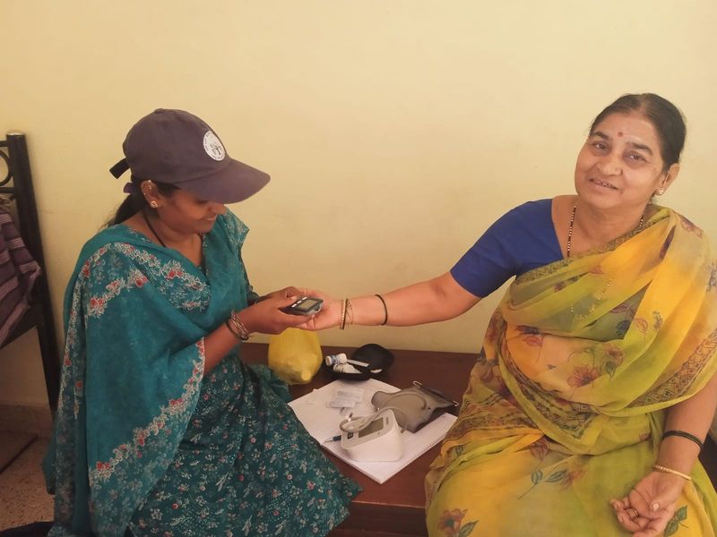 A community health volunteer checks a smiling woman's blood sugar at her home.