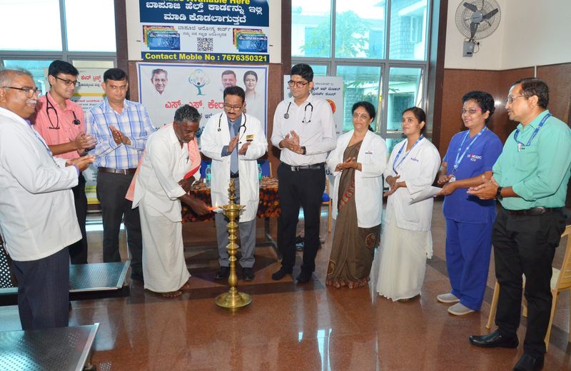 Doctors and staff in white coats watch an older man light a ceremonial brass lamp to open a health programme, below an SS Care Trust banner.
