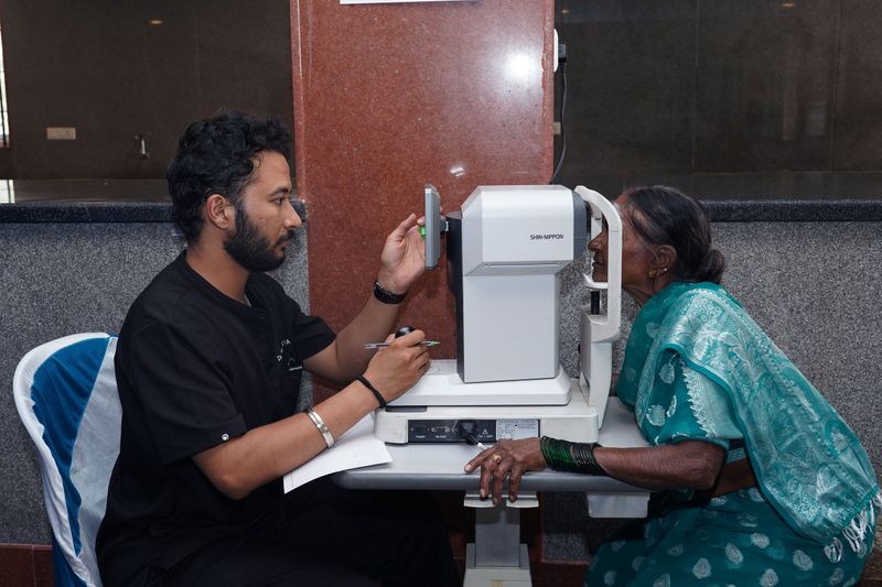 An optometrist operates an eye-testing machine while an older woman in a teal saree rests her chin on it for an eye screening.