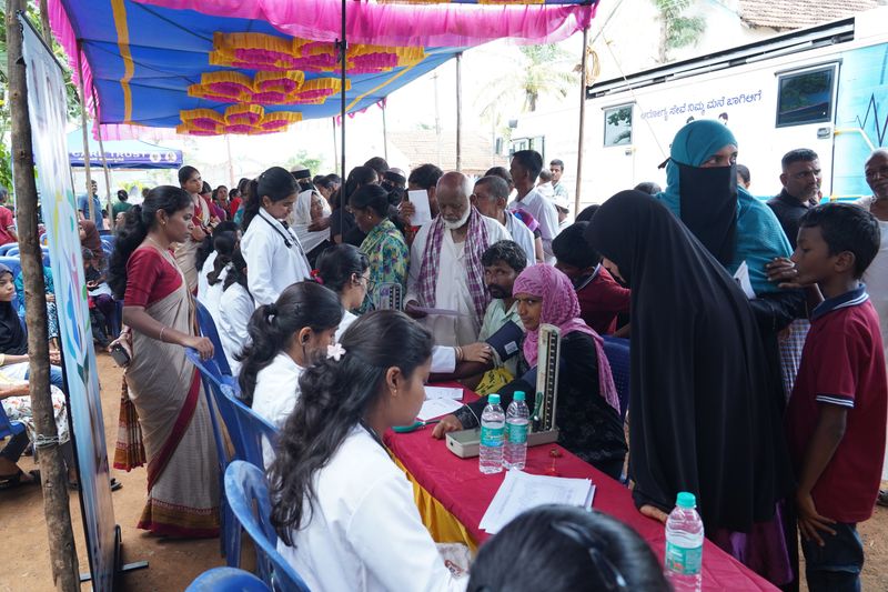 Residents queue for blood-pressure checks under a canopy beside the SS Care Trust Health-on-Wheels mobile clinic van.