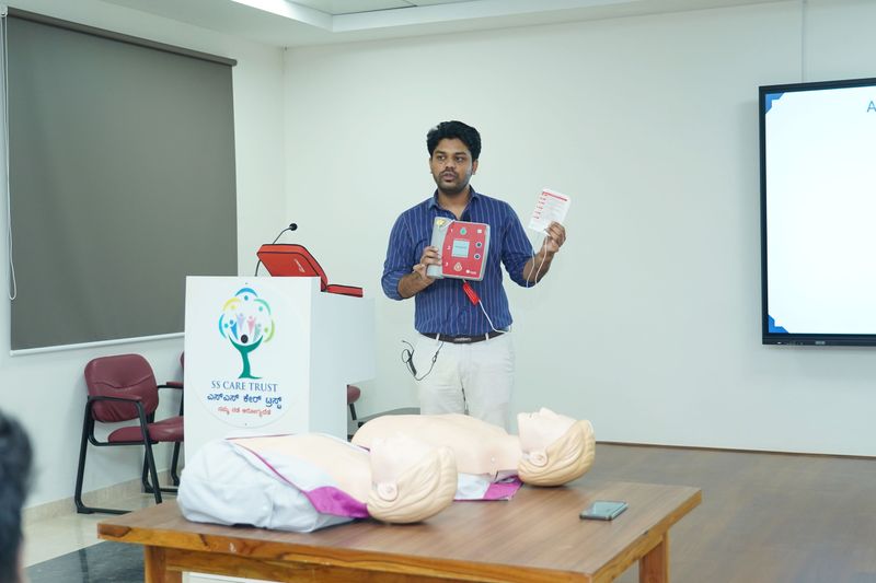 A facilitator demonstrates an AED device beside a CPR training manikin at an SS Care Trust session.