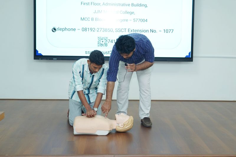 Two men practice hands-on chest compressions on a CPR training manikin on the floor.