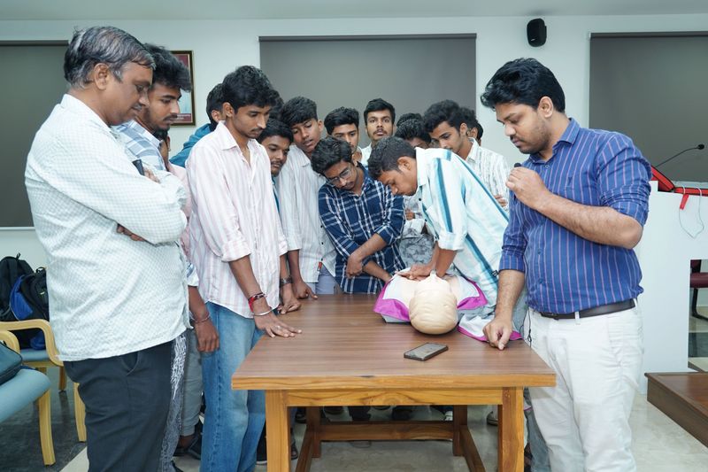 A group of young men gather around a table to observe a chest-compression demonstration on a manikin.