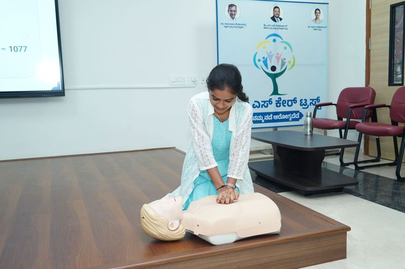 A woman kneels and performs chest compressions on a CPR manikin during a hands-on training exercise.