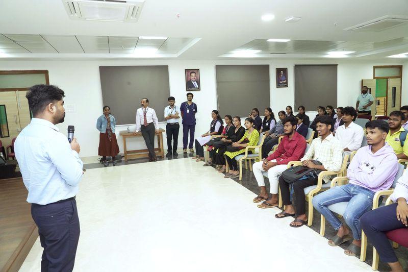 A facilitator addresses a seated audience in a hall with portrait photos on the wall during CPR training.