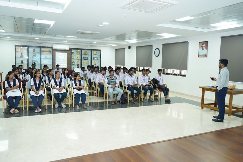 A trainer addresses rows of seated students, including girls in school uniform, during a first aid session.