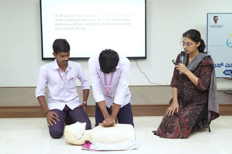 Two trainees practice chest compressions on a manikin while a facilitator explains an emergency scenario.