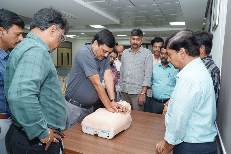 A group of men watches closely as a facilitator demonstrates chest compressions on a CPR manikin.