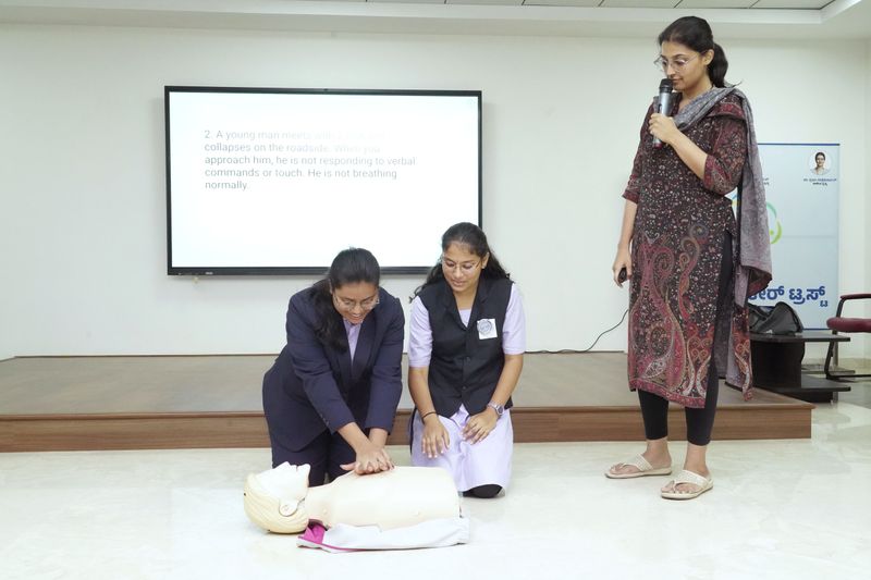 Two young women practice chest compressions on a manikin as a facilitator looks on.