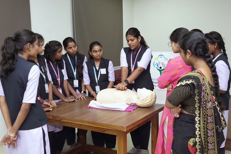 A group of schoolgirls gathers around a table to watch a classmate practice CPR on a training manikin.