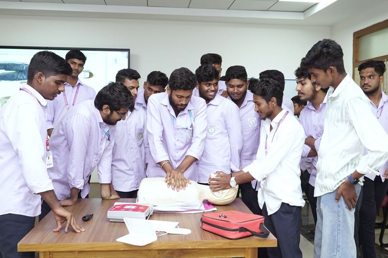 A group of young men in uniform shirts gather closely around a training mannequin as an instructor demonstrates CPR chest compressions.
