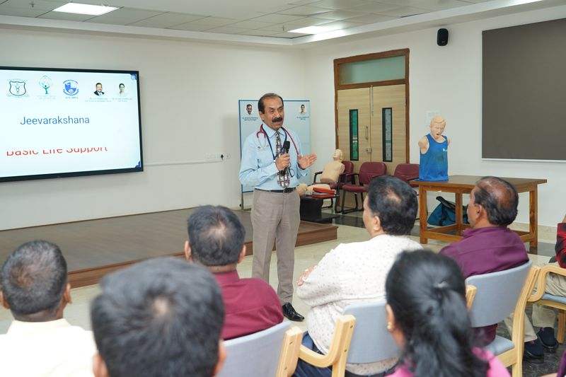A trainer addresses a seated audience under a screen titled 'Jeevarakshana: Basic Life Support,' with training mannequins set up nearby.