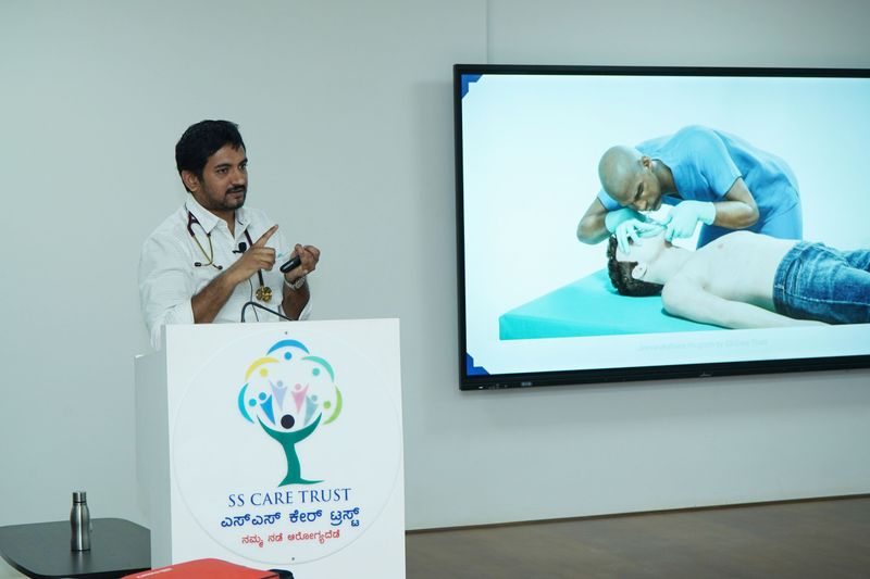 A trainer speaks from an SS Care Trust podium beside a screen showing a demonstration photo of an airway-opening first aid technique.