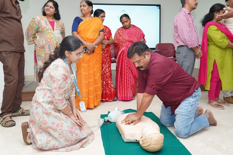 A man practices chest compressions on a training mannequin while women from a community group observe a hands-on first aid session.