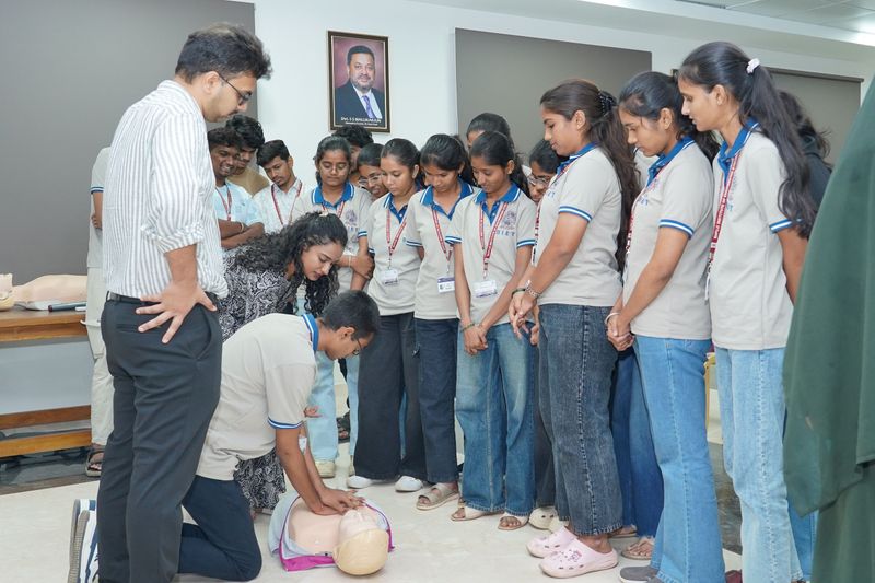 An instructor kneels to demonstrate CPR chest compressions on a mannequin as a group of attentive students watches closely.