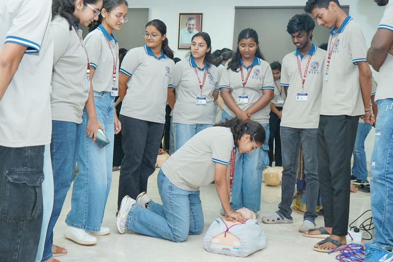 Engineering college student practising chest compressions on a training manikin while classmates look on during a first aid session.