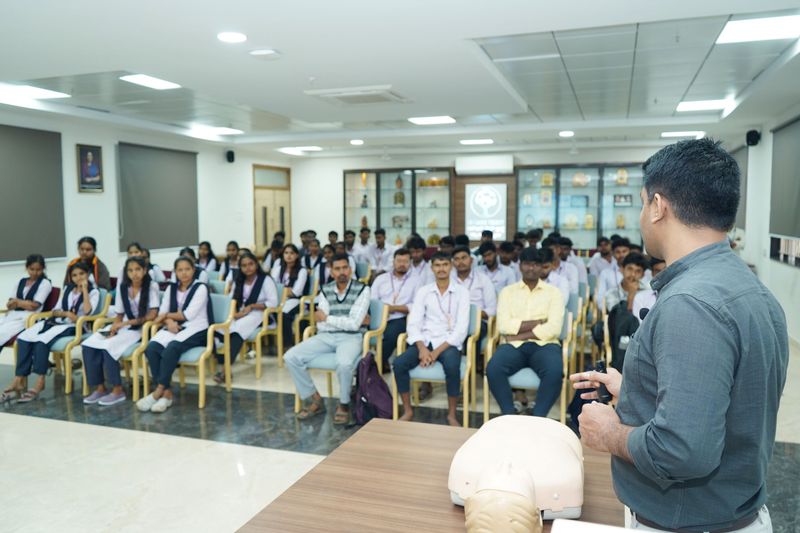 Students in a lecture hall watching a trainer demonstrate CPR technique with a training manikin.