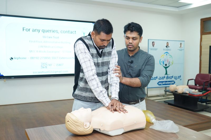 Two men practising chest compressions on a CPR manikin on a table during a first aid training session.