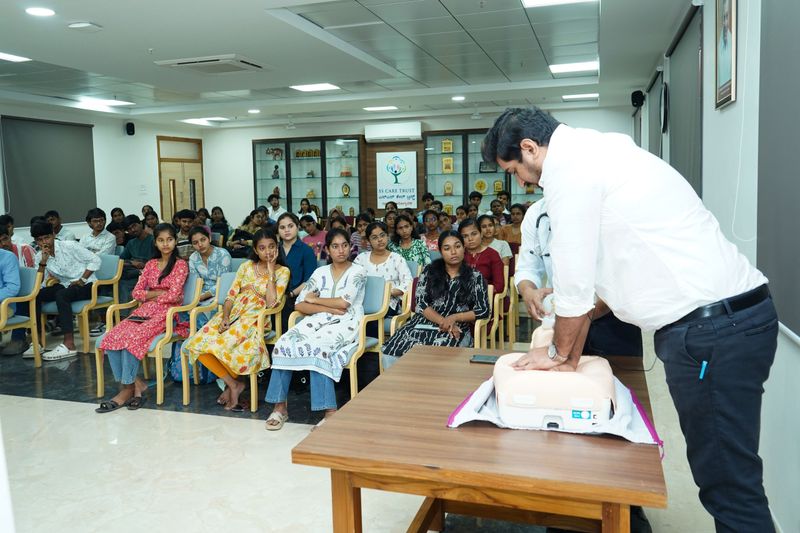 Trainer demonstrating CPR technique on a manikin to a seated audience of women in colourful attire.