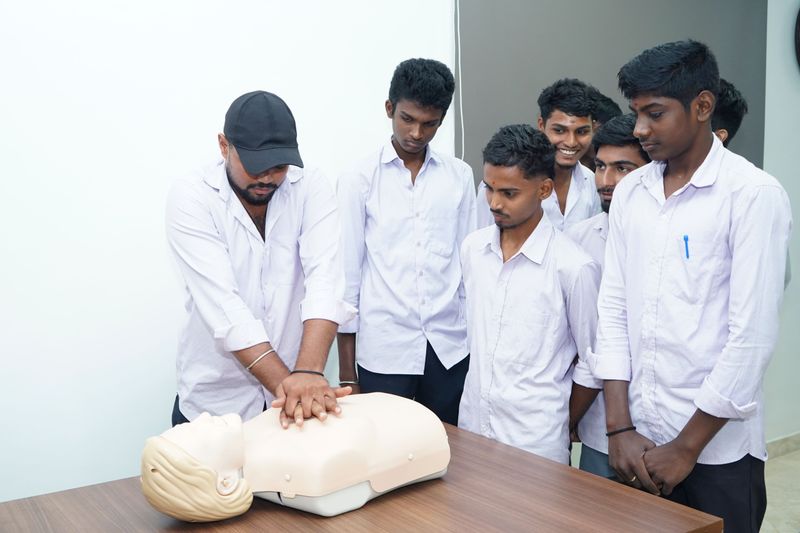 Trainer wearing a cap demonstrating chest compressions on a manikin to a group of young men.