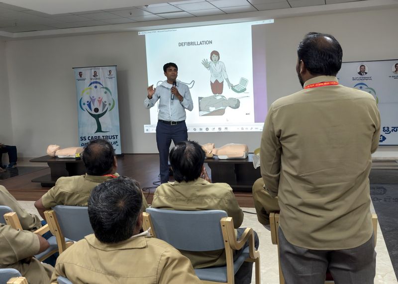 A trainer explains defibrillation using a projected slide to seated uniformed workers at an SS Care Trust first aid session, with CPR manikins laid out nearby.