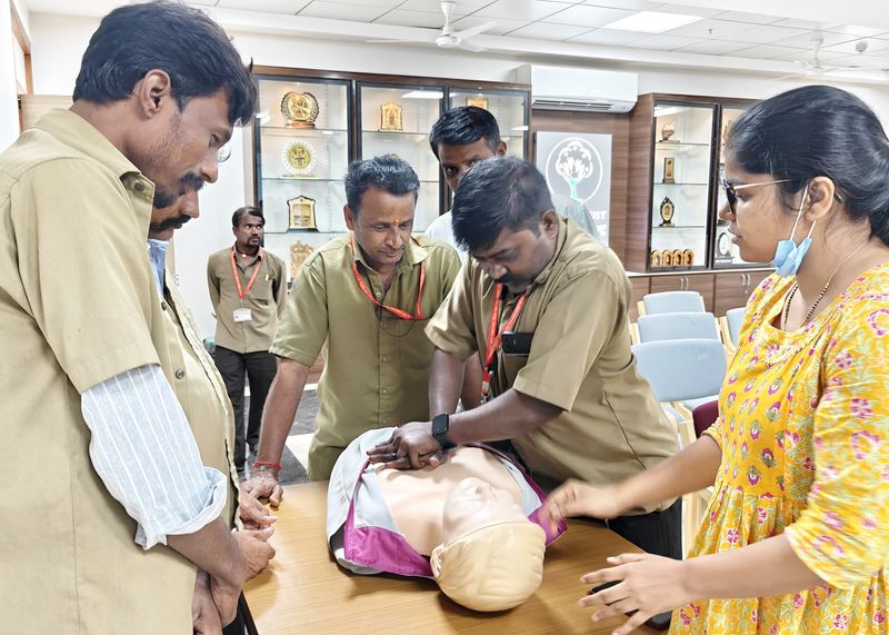 Trainees practice chest compressions on a CPR manikin at a table while classmates and a facilitator look on.