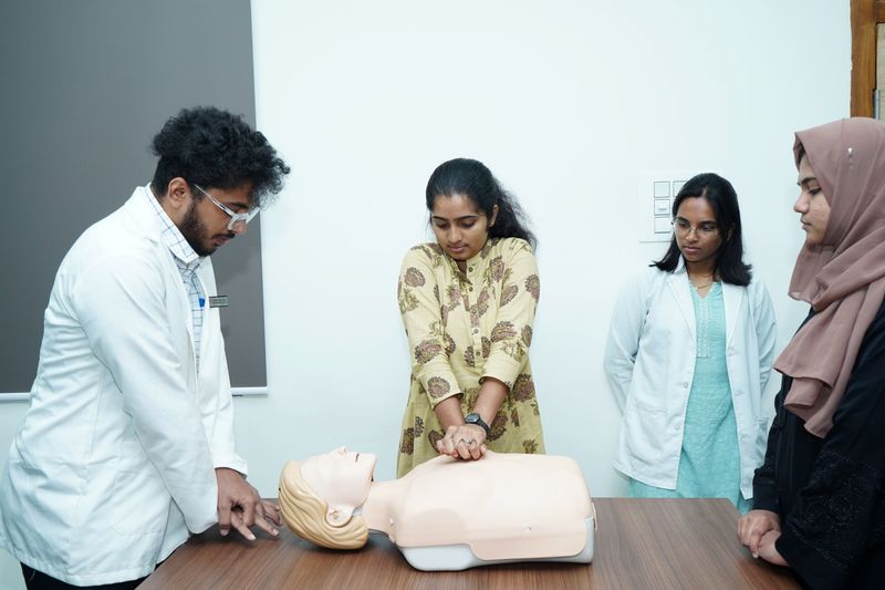A trainee practices chest compressions on a CPR manikin at a table while three colleagues, two in white coats, observe closely.