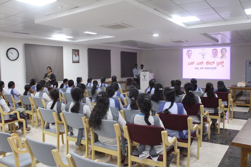 Rows of schoolgirls in uniform listen attentively during an SS Care Trust health awareness session, with the trust's logo on screen.