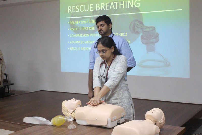 Two facilitators demonstrate chest compressions on a CPR manikin beneath a projected 'Rescue Breathing' training slide.