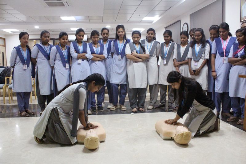 Two schoolgirls kneel and practice chest compressions on CPR manikins while classmates in uniform watch attentively.