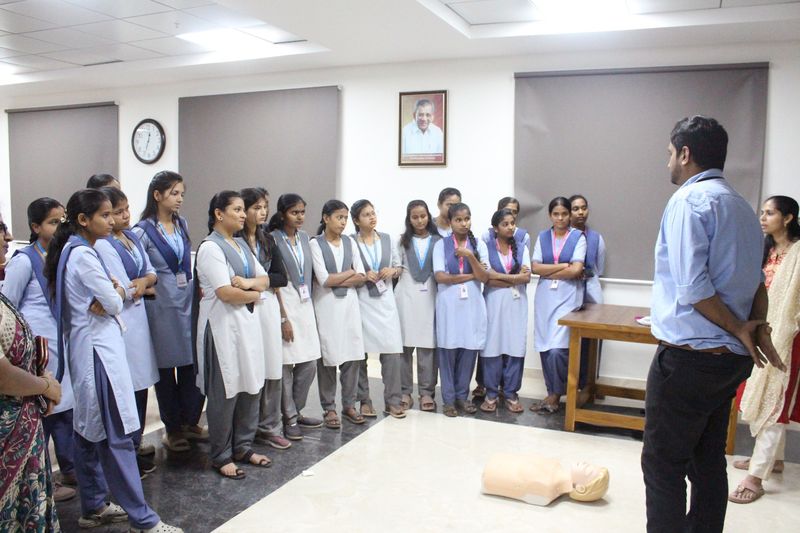 A facilitator addresses a line of schoolgirls standing beside a CPR manikin placed on the floor during a life support session.