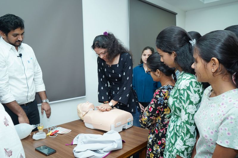 A facilitator demonstrates chest compressions on a manikin to a small group of young women during a training session.