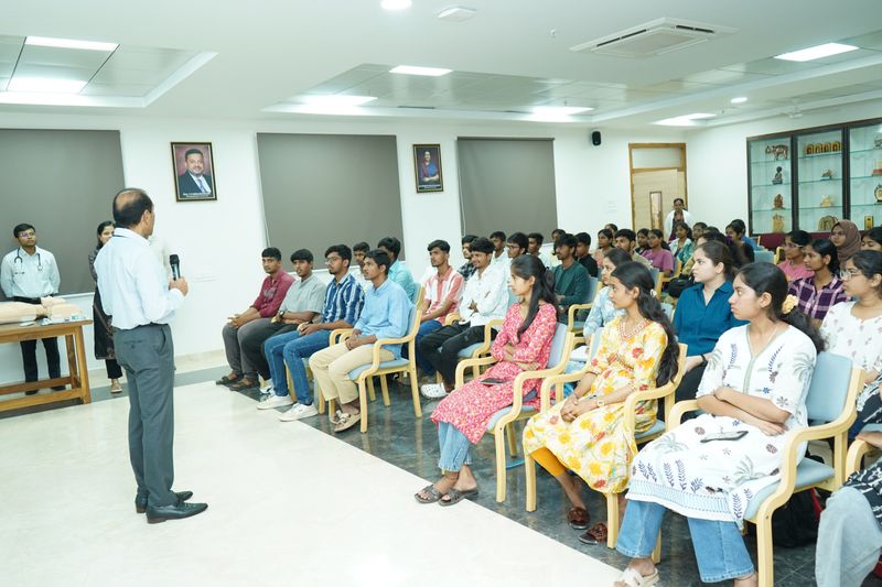 A facilitator addresses a seated audience in a hall, with a CPR manikin placed on a table beside him during a first aid training.