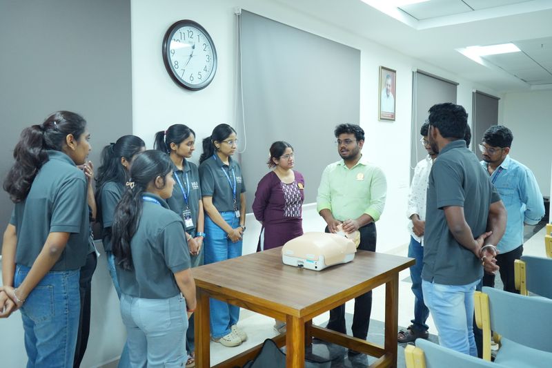 A facilitator demonstrates CPR on a manikin at a table while a group of uniformed trainees gather around to observe.
