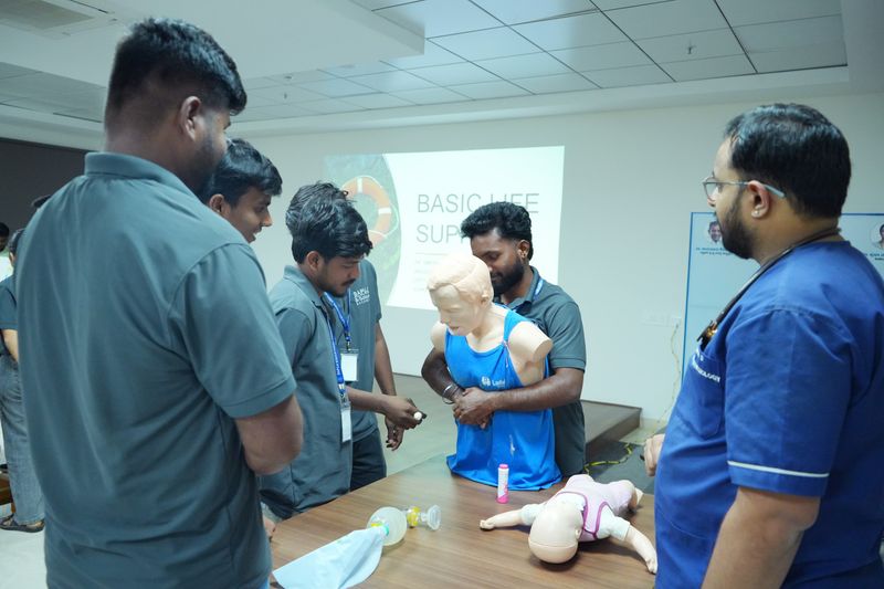 A facilitator demonstrates CPR on a child-sized manikin while trainees look on, with a 'Basic Life Support' slide projected behind them.