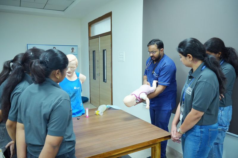 A facilitator holds an infant CPR manikin to demonstrate technique to a group of trainees gathered around a table.