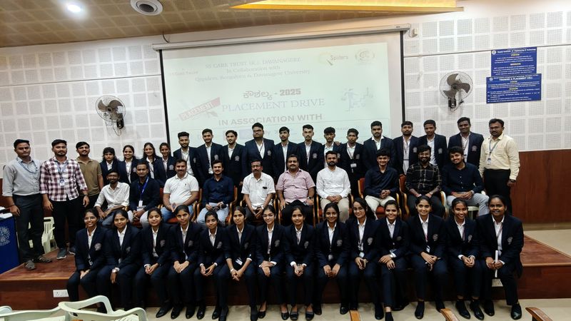 About forty students in dark blazers pose with organisers in front of a projected Kaushalya 2025 Placement Drive slide naming SS Care Trust, Qspiders and Davangere University.