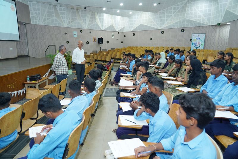 Rural school students in blue uniforms sit with notebooks as two facilitators lead a Kaushalya session in an auditorium.