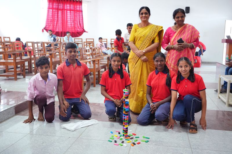 Five students pose with two women facilitators beside a colourful block tower built during a Kaushalya activity.
