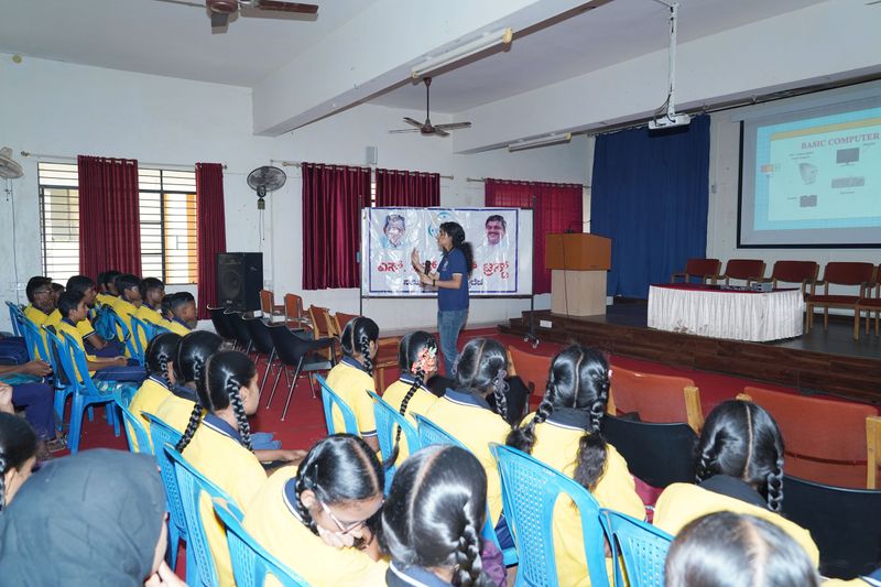 Students sit in a hall as a facilitator leads a basic computer lesson shown on the projector screen.
