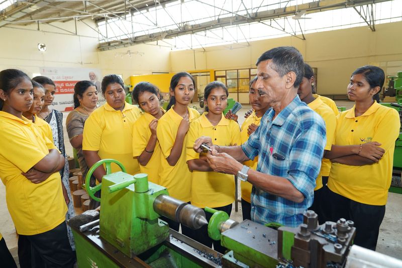 A group of students watches an instructor explain a machine part during a Kaushalya workshop visit.