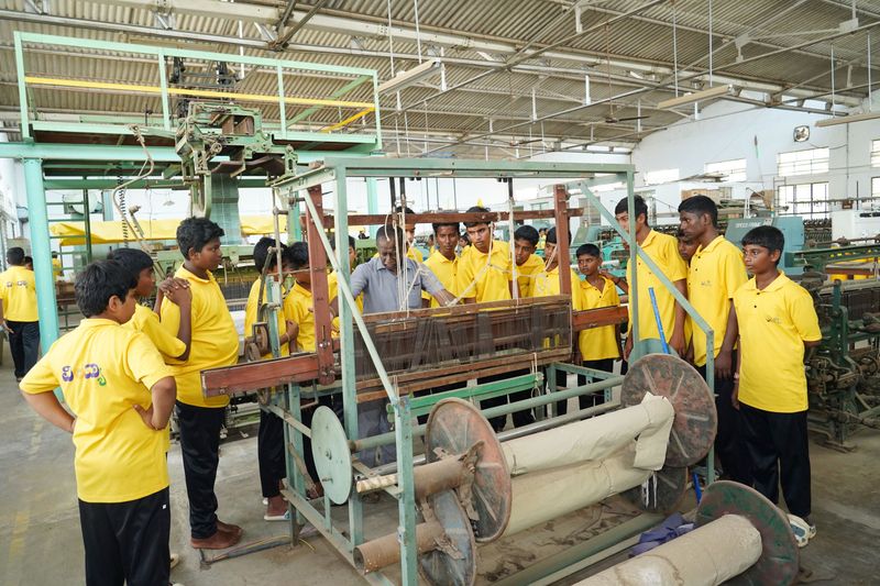 Students in yellow uniform shirts gather around a handloom machine as an instructor explains its parts during a skill-building visit.