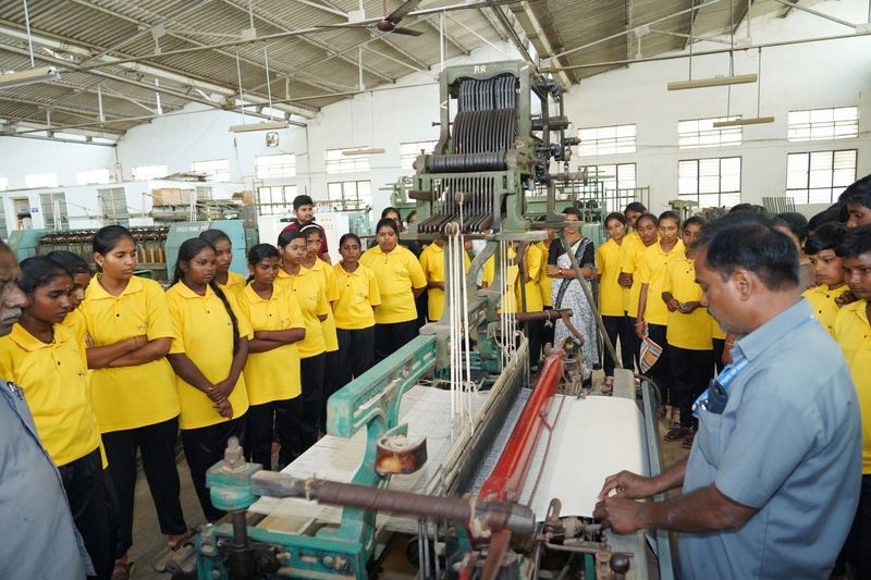 A group of students in yellow uniforms watch closely as a staff member demonstrates a handloom machine in a workshop hall.