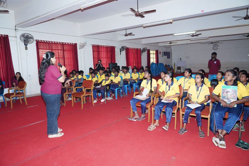 A facilitator holding a microphone addresses a hall of seated students during an interactive session.