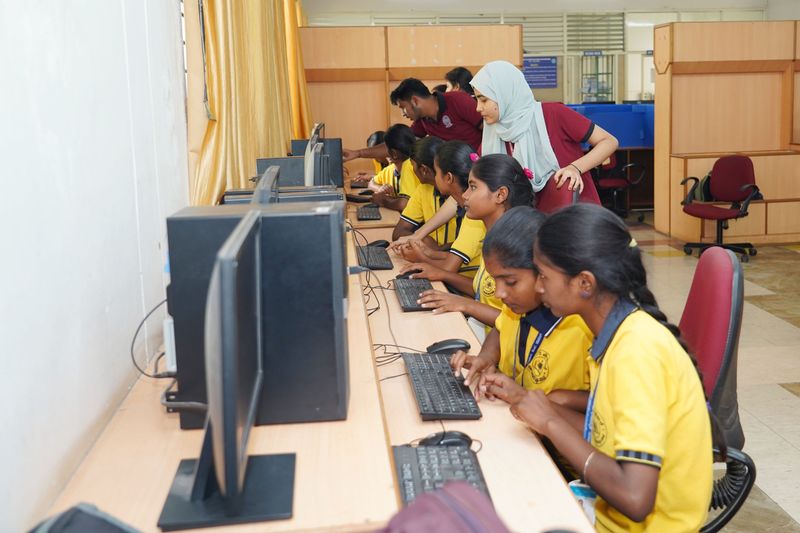 Students in yellow uniforms practice on desktop computers in a lab while two mentors guide them.