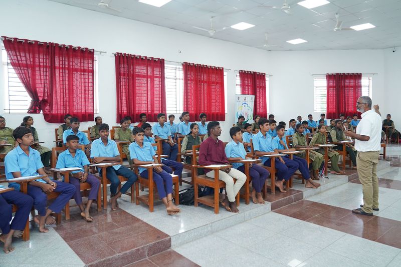 A speaker addresses rows of seated school students in blue and olive uniforms during a classroom talk.