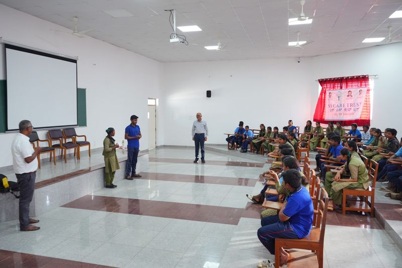 Wide view of a hall with students and staff seated for a session, with an S S Care Trust banner visible on the back wall.