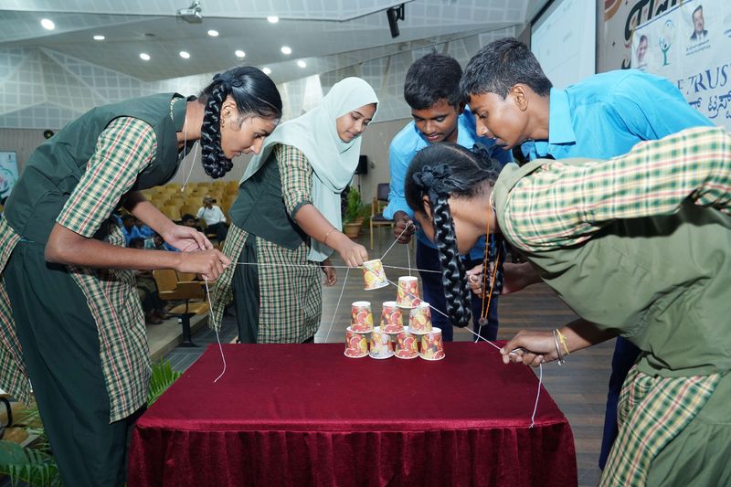 Students work together pulling strings to stack cups into a pyramid during a team-building exercise.